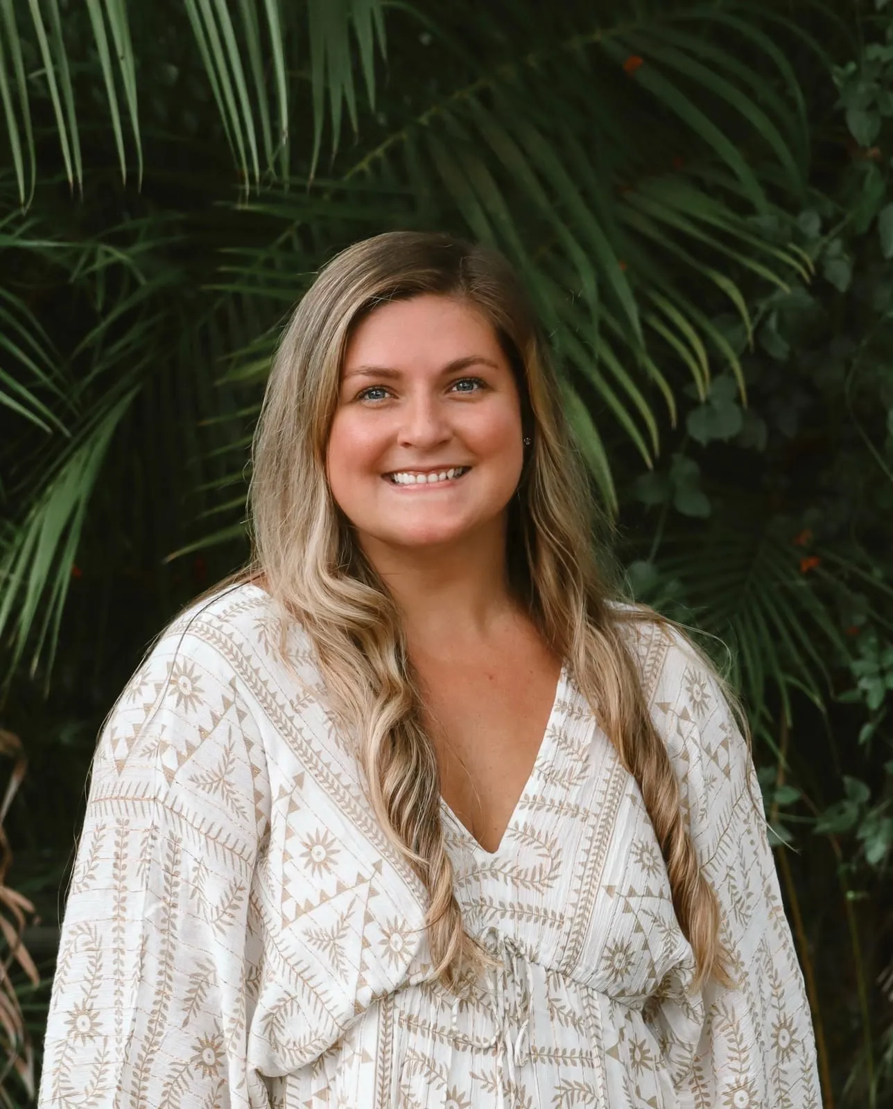 Mel Headshot Smiling woman with long hair in patterned dress, surrounded by lush greenery.