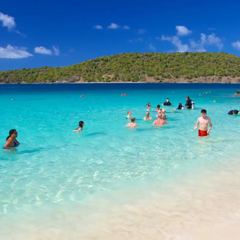 People enjoying a sunny day at a beach with clear blue water and green hills in the background.