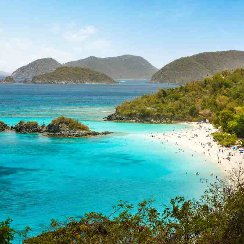 Tropical beach with turquoise water, lush hills, and distant islands under a clear blue sky.