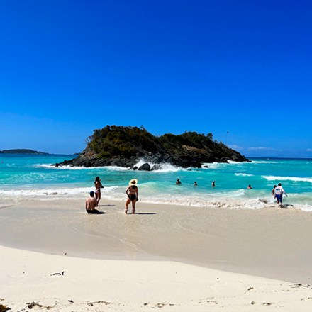 People enjoying a sunny day at a beach with clear water and a distant island.