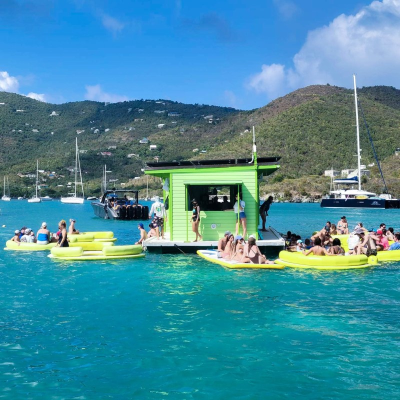 People on floating dock with green hut and inflatable rings in a bay with sailboats and hills.