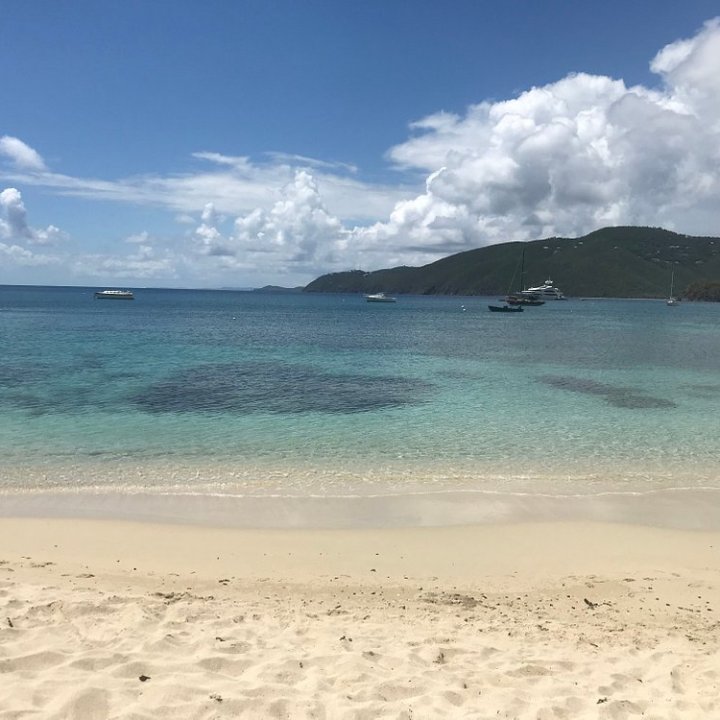 Sandy beach with clear blue water and boats under a partly cloudy sky.