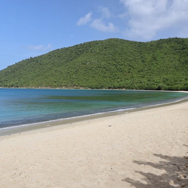 Sandy beach with clear blue water and green hills under a partly cloudy sky.