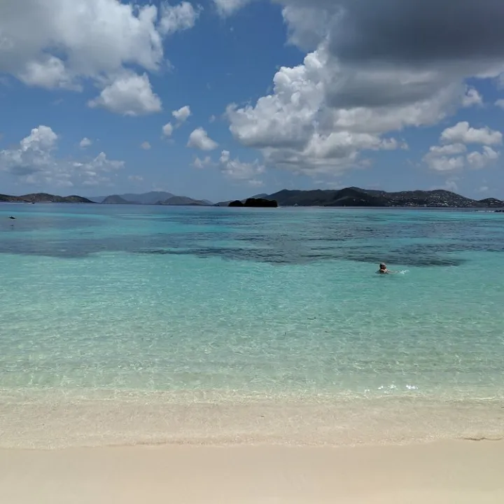 Person swimming in clear turquoise ocean under partly cloudy sky with distant hills.