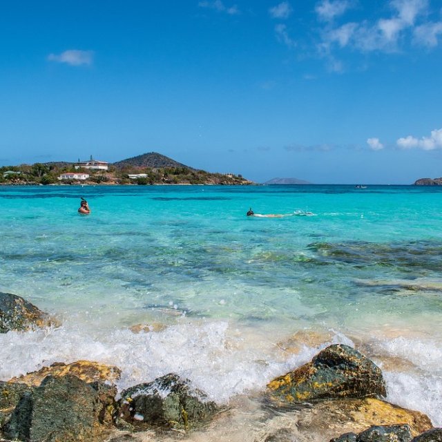 Beach with turquoise water, rocks, and distant hills under a clear blue sky; people swimming.