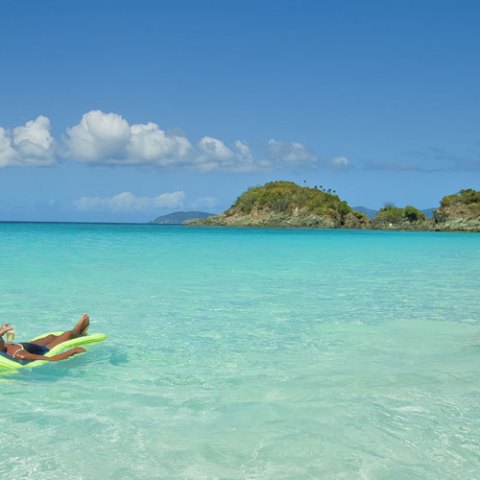 Person relaxing on a float in clear turquoise water with small islands and blue sky in background.
