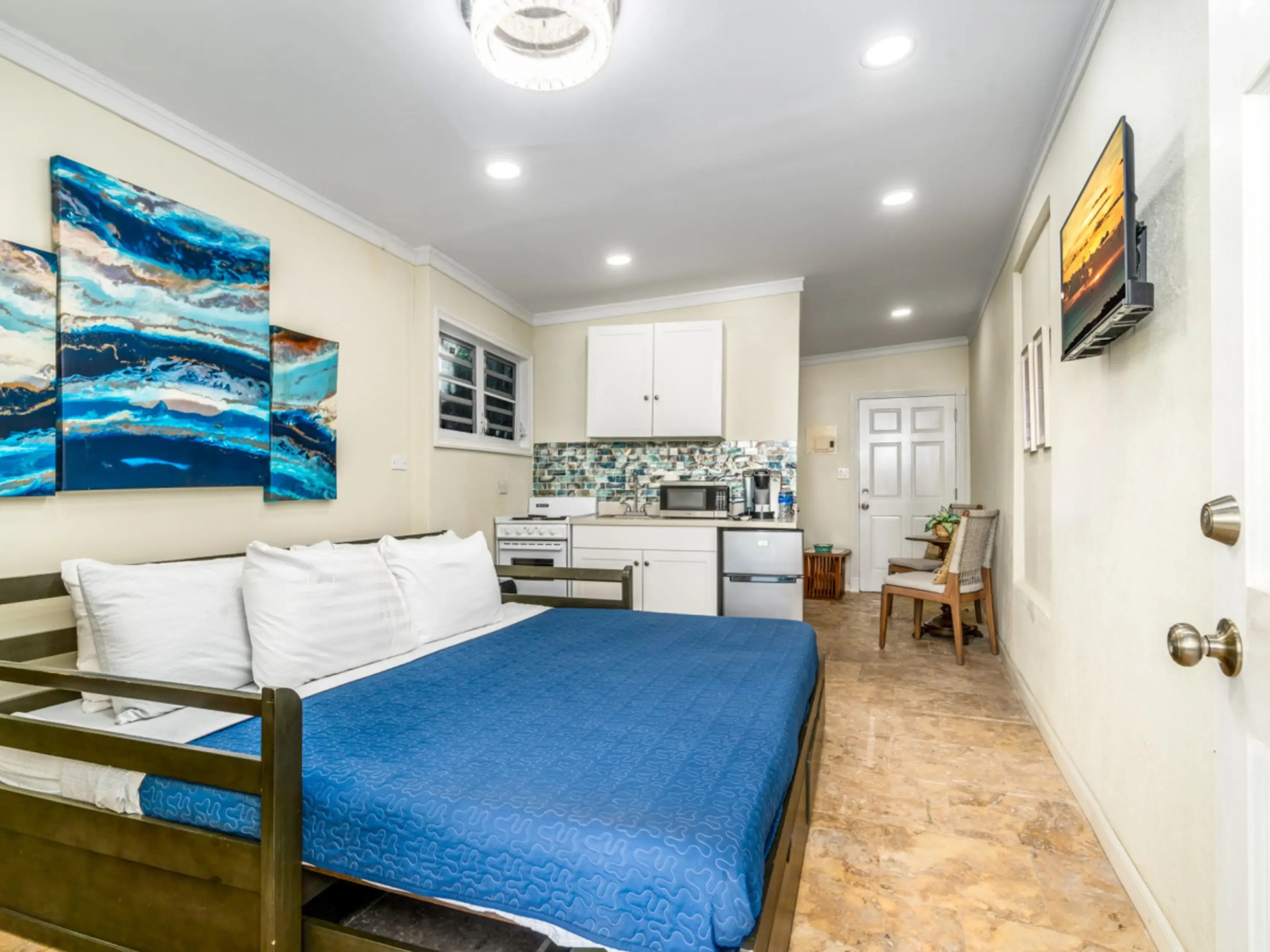 Room with blue bedspread, abstract art, kitchen area, and wall-mounted TV.