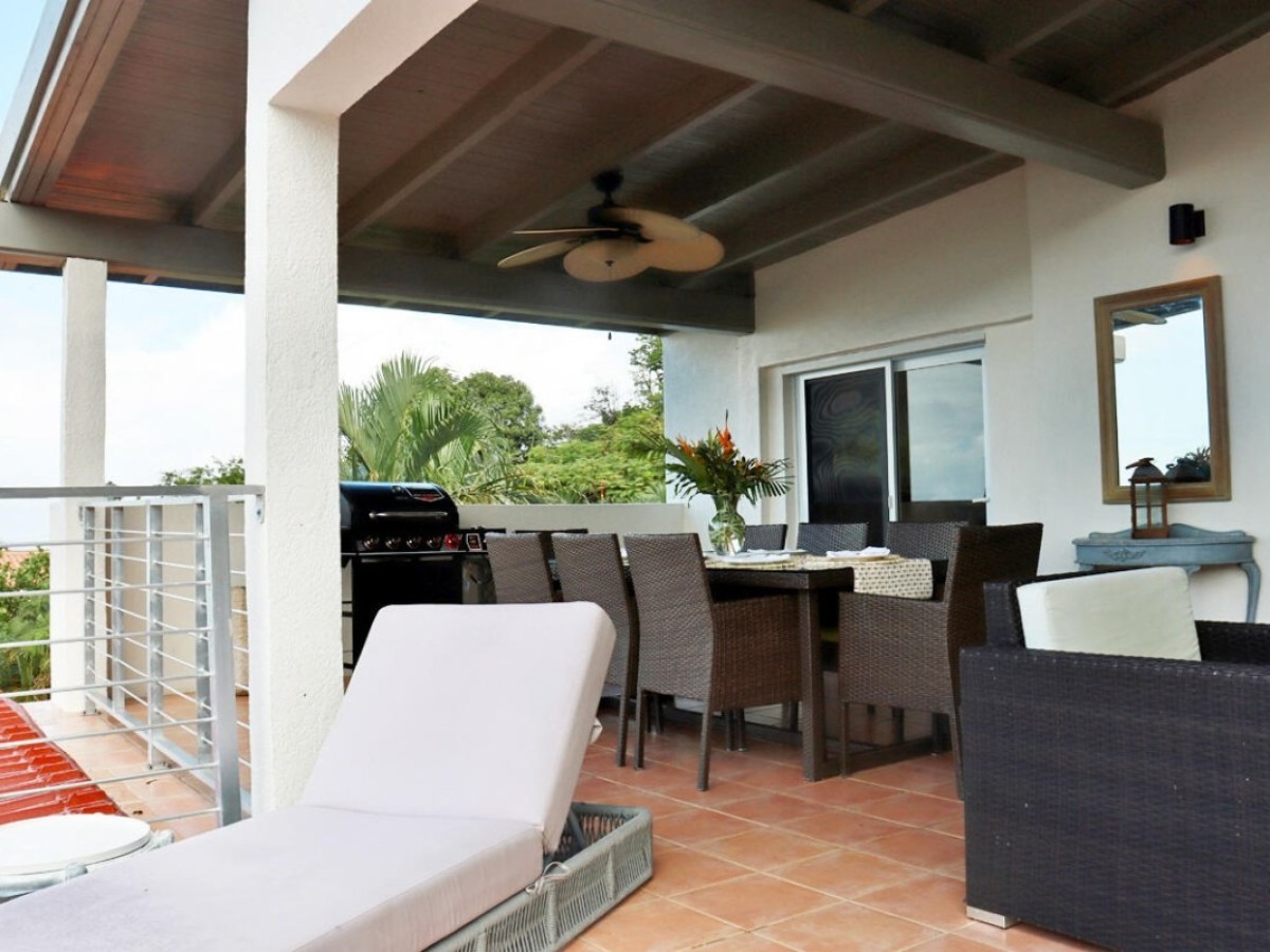 Covered patio with dining table, chairs, ceiling fan, and lounge chair on tiled floor.