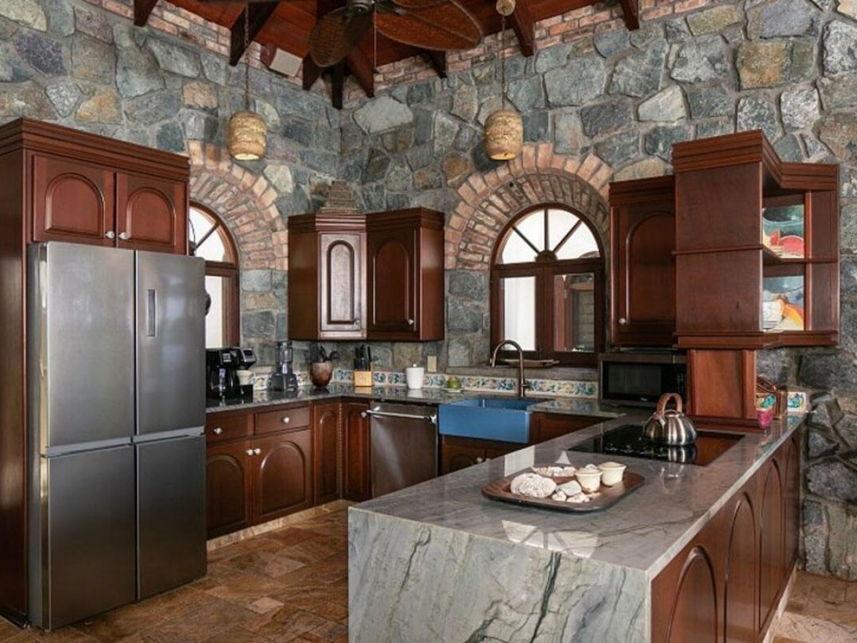 Stone-walled kitchen with wooden cabinets and marble island, featuring stainless fridge and arched windows.