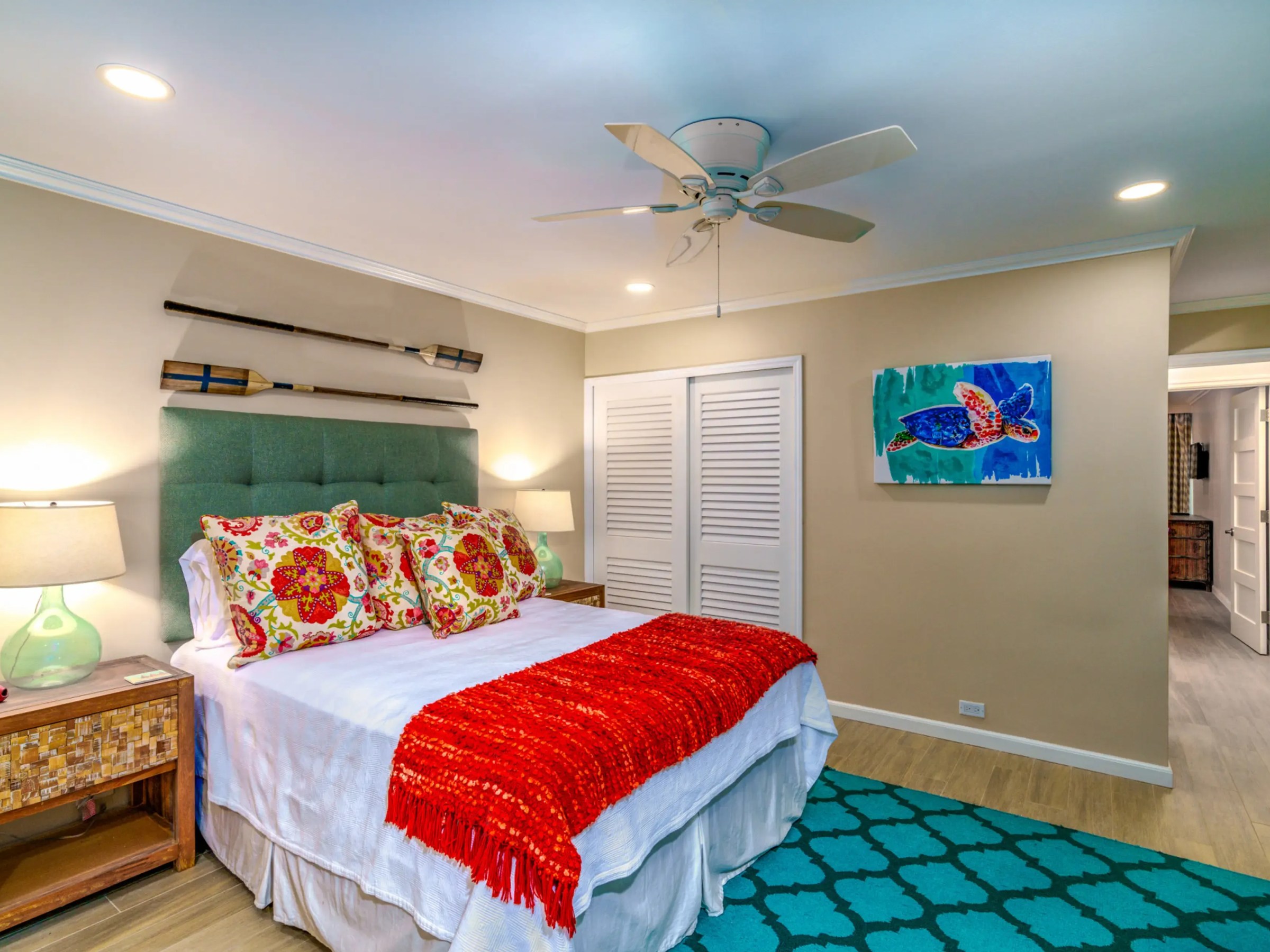Bedroom with a bed, colorful cushions, fan, art on the wall, and a blue rug.