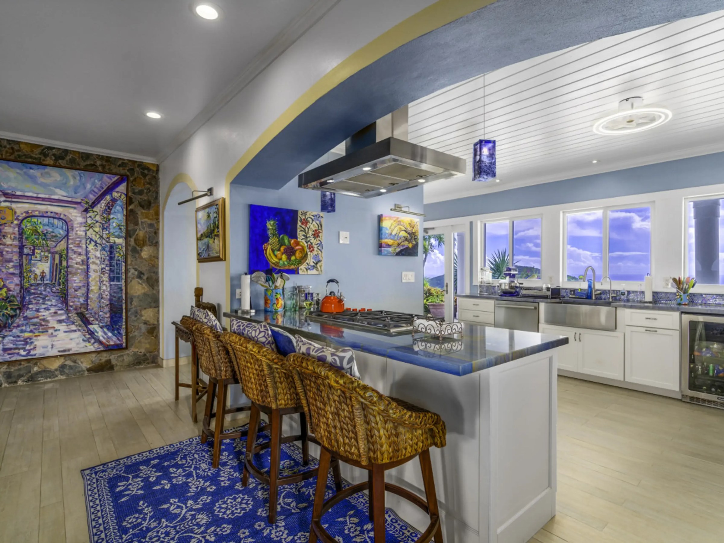 Modern kitchen with island, bar stools, large painting on wall, and windows showing ocean view.