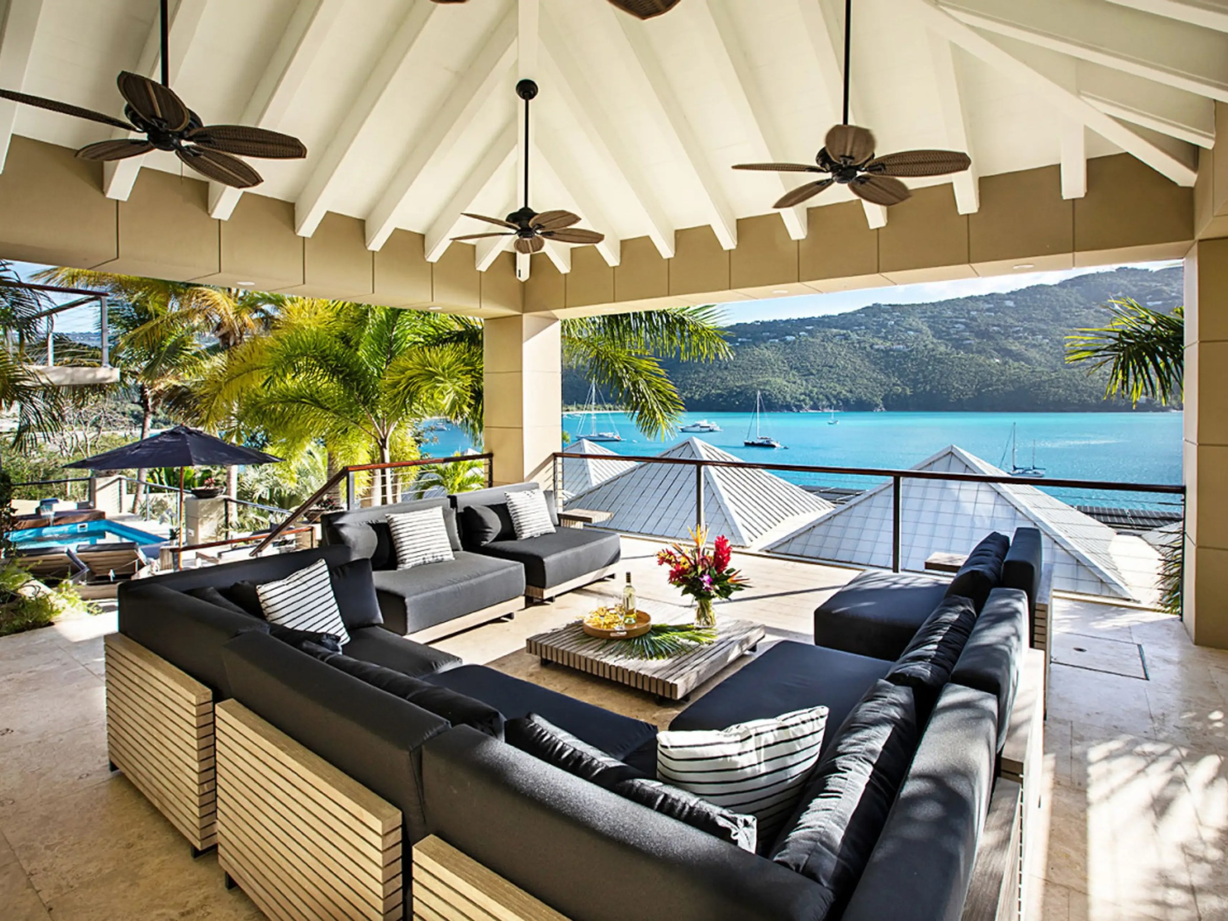 Open patio with black couches, ceiling fans, and ocean view through palm trees.