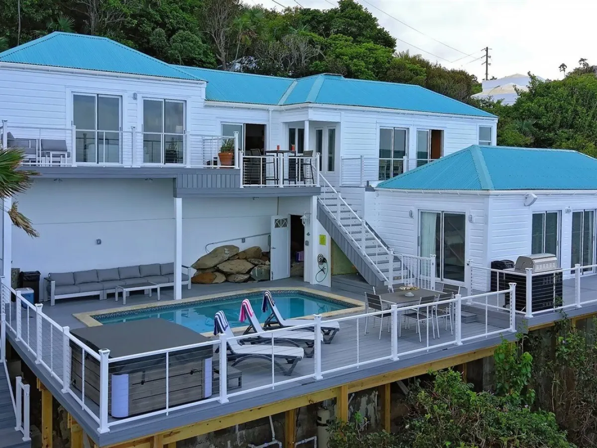 White house with blue roof, elevated deck, pool, patio furniture, and surrounded by trees.