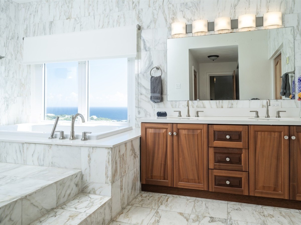 Luxurious bathroom with marble tub, ocean view window, and wooden vanity under large mirror.