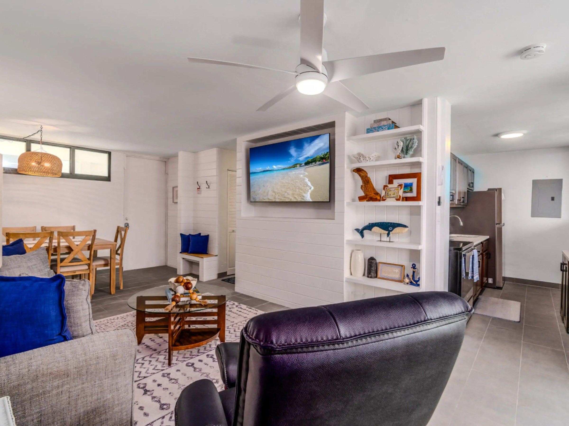 Modern living room with gray sofa, blue pillows, dining table, TV, ceiling fan, and kitchen in open layout.