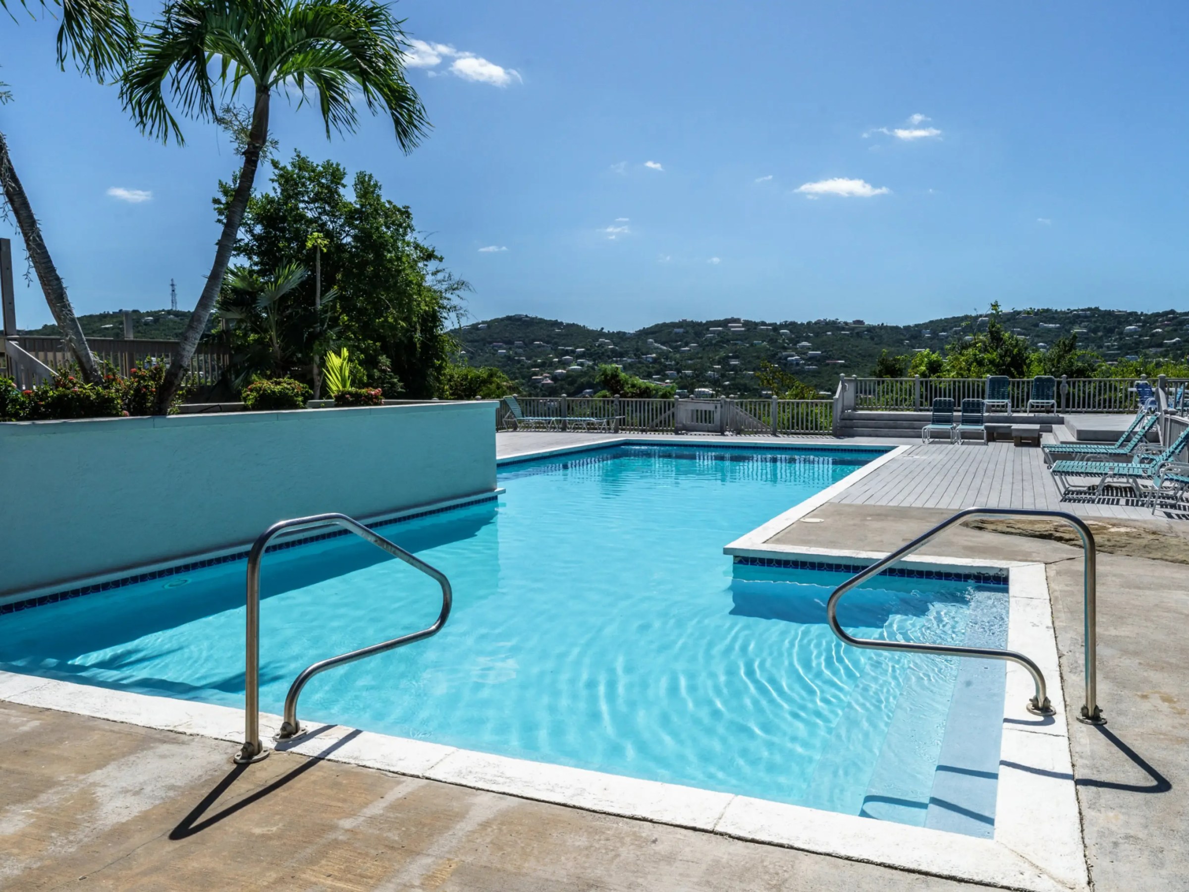 Outdoor pool with metal handrails, surrounded by trees and hills under a clear blue sky.