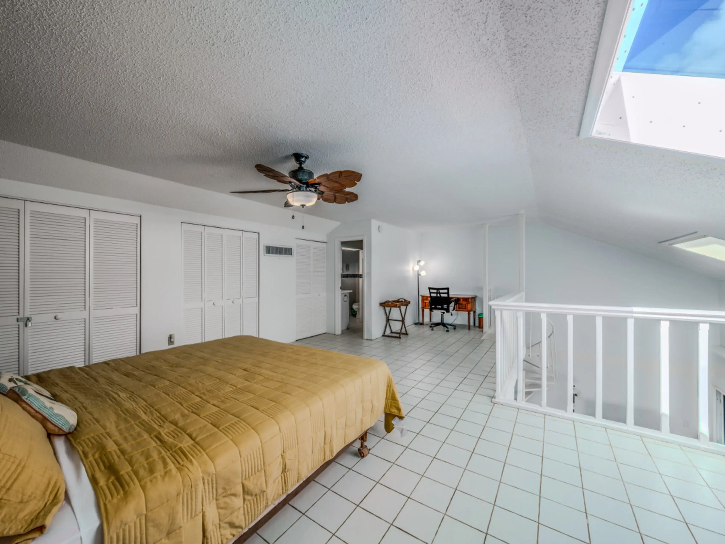 Spacious bedroom with beige bedspread, ceiling fan, skylight, and white tiled floor.