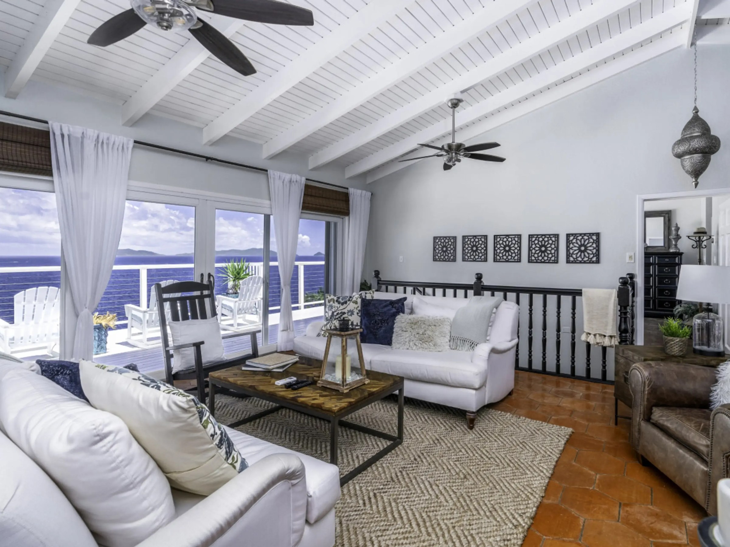 Coastal living room with white sofas, ceiling fans, and ocean view through large windows.