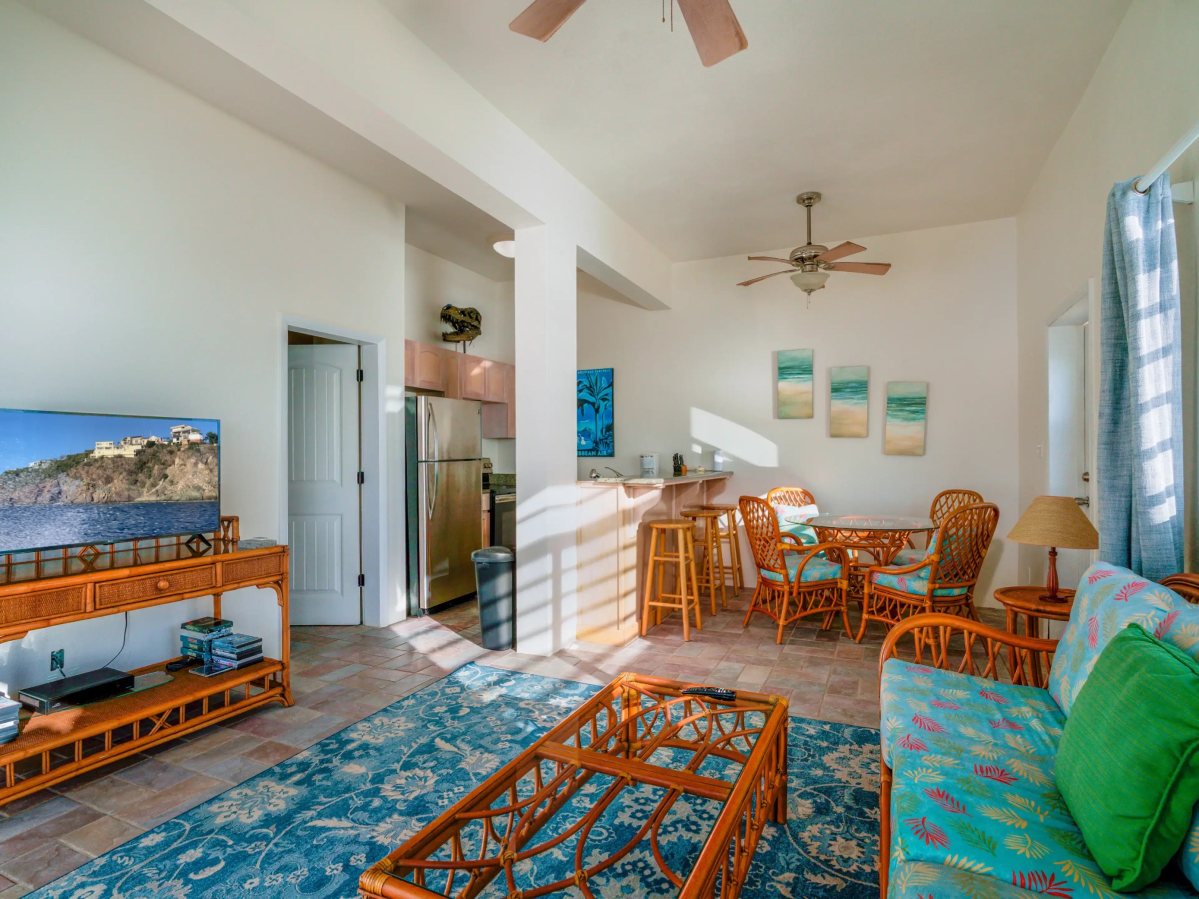 Beach-themed living room with wicker furniture, TV, and decor, featuring a kitchen area and ceiling fan.