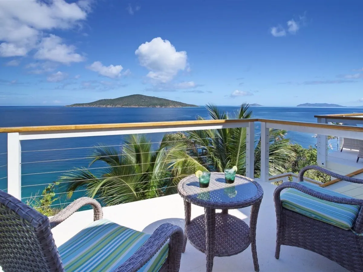 Patio with wicker chairs overlooks ocean and small island, clear skies with few clouds.