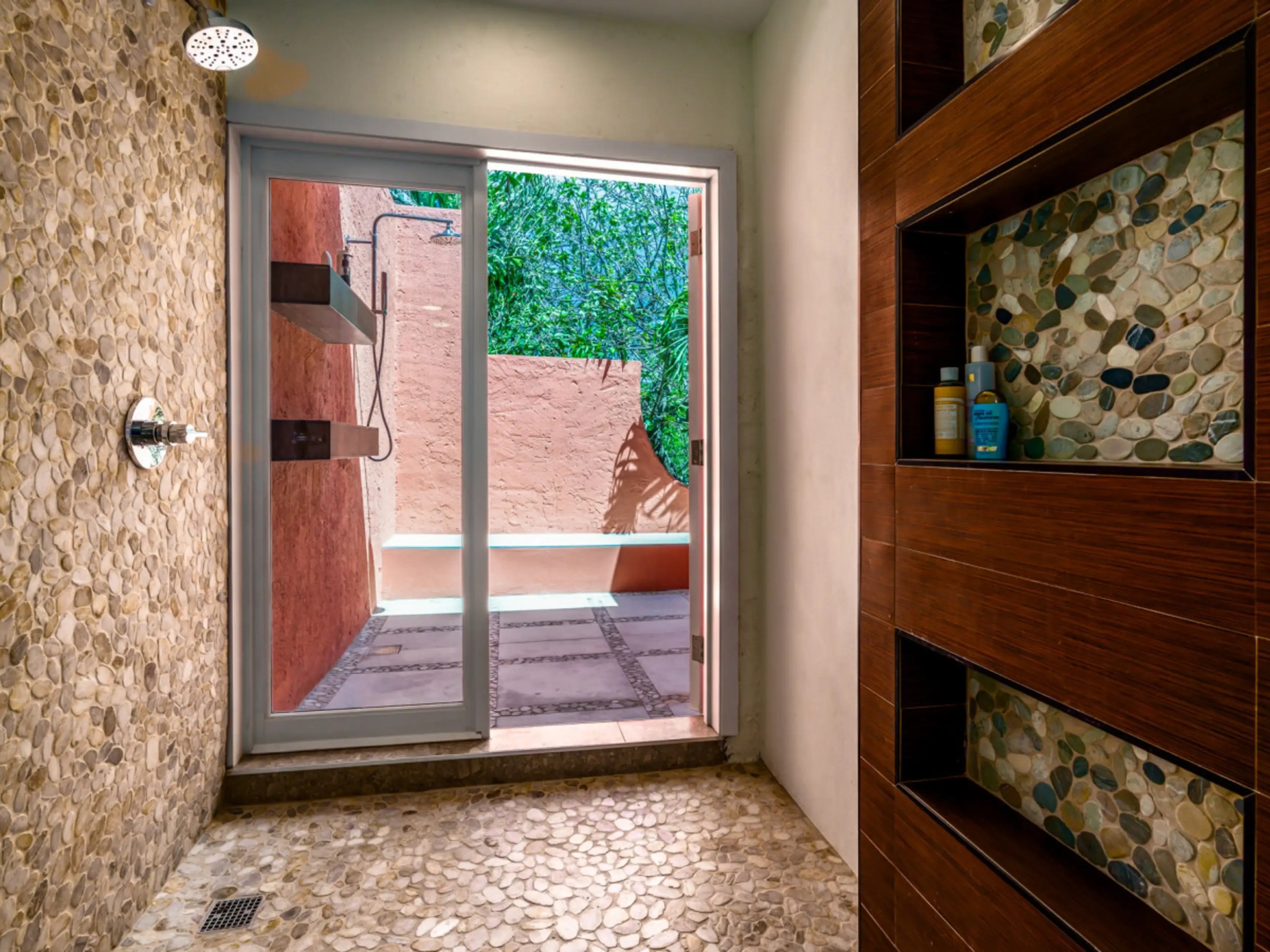 Stone-tiled shower with glass door opening to outdoor patio with greenery.