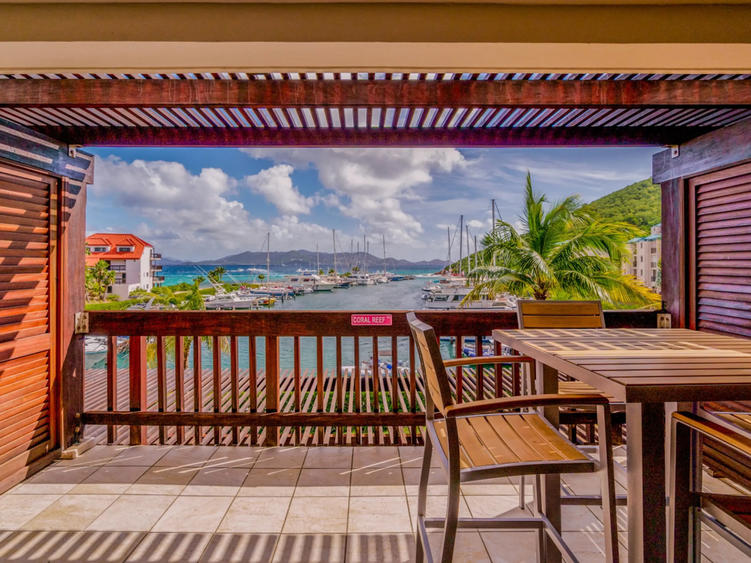 Wooden patio with table and chairs overlooking a marina with boats and palm trees.