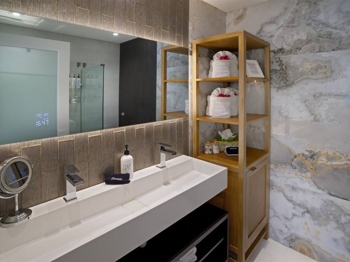 Modern bathroom with double sink, marble wall, large mirror, and wooden shelf with towels.
