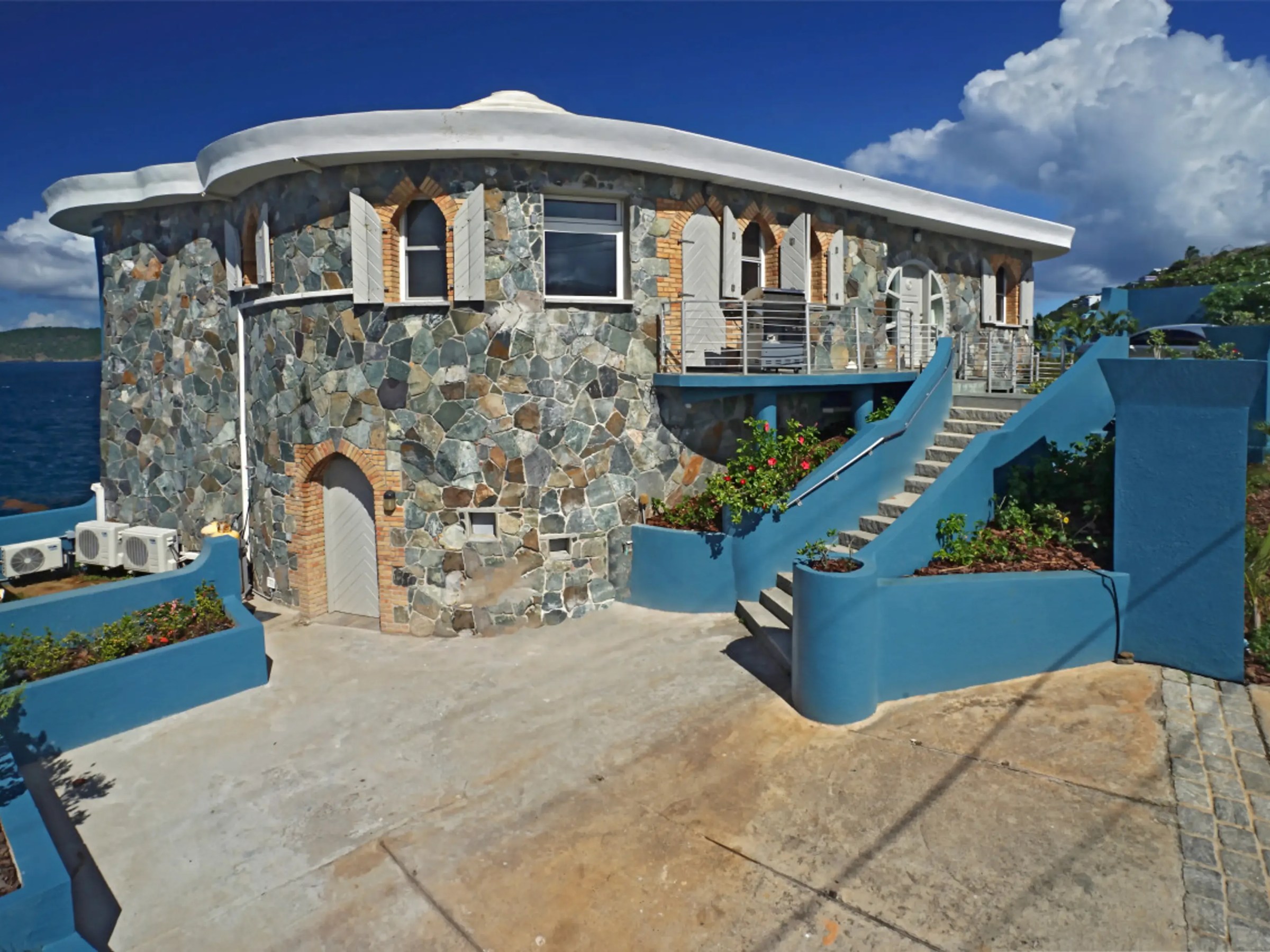 Stone house with round tower, blue trim, by the sea under a clear blue sky.