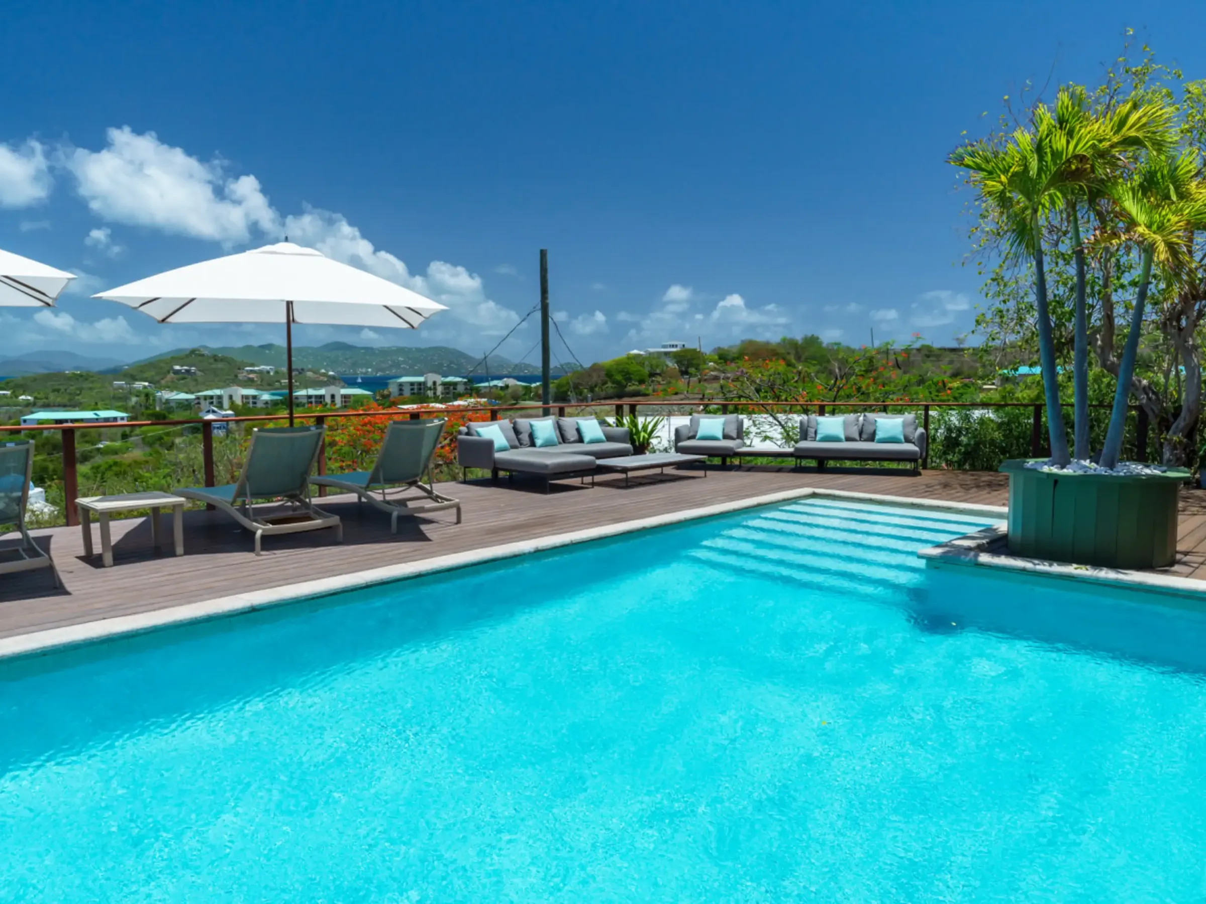 Outdoor pool with loungers, umbrellas, and distant view; tropical setting with clear sky.