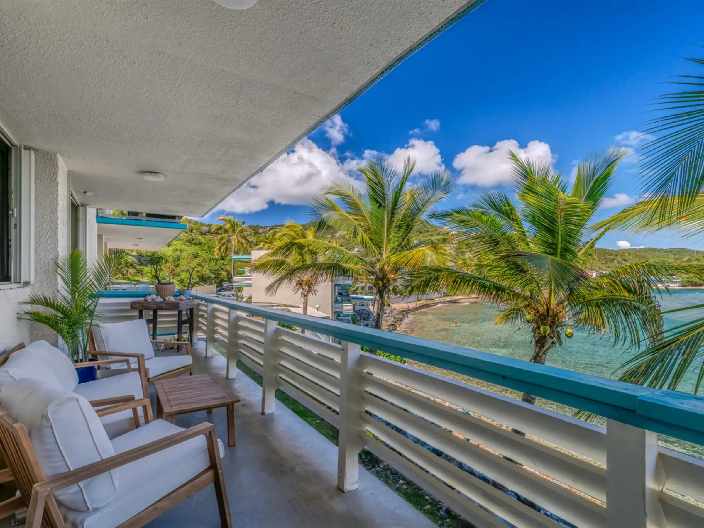 Balcony with white chairs and table, overlooking tropical beach with palm trees and blue sky.