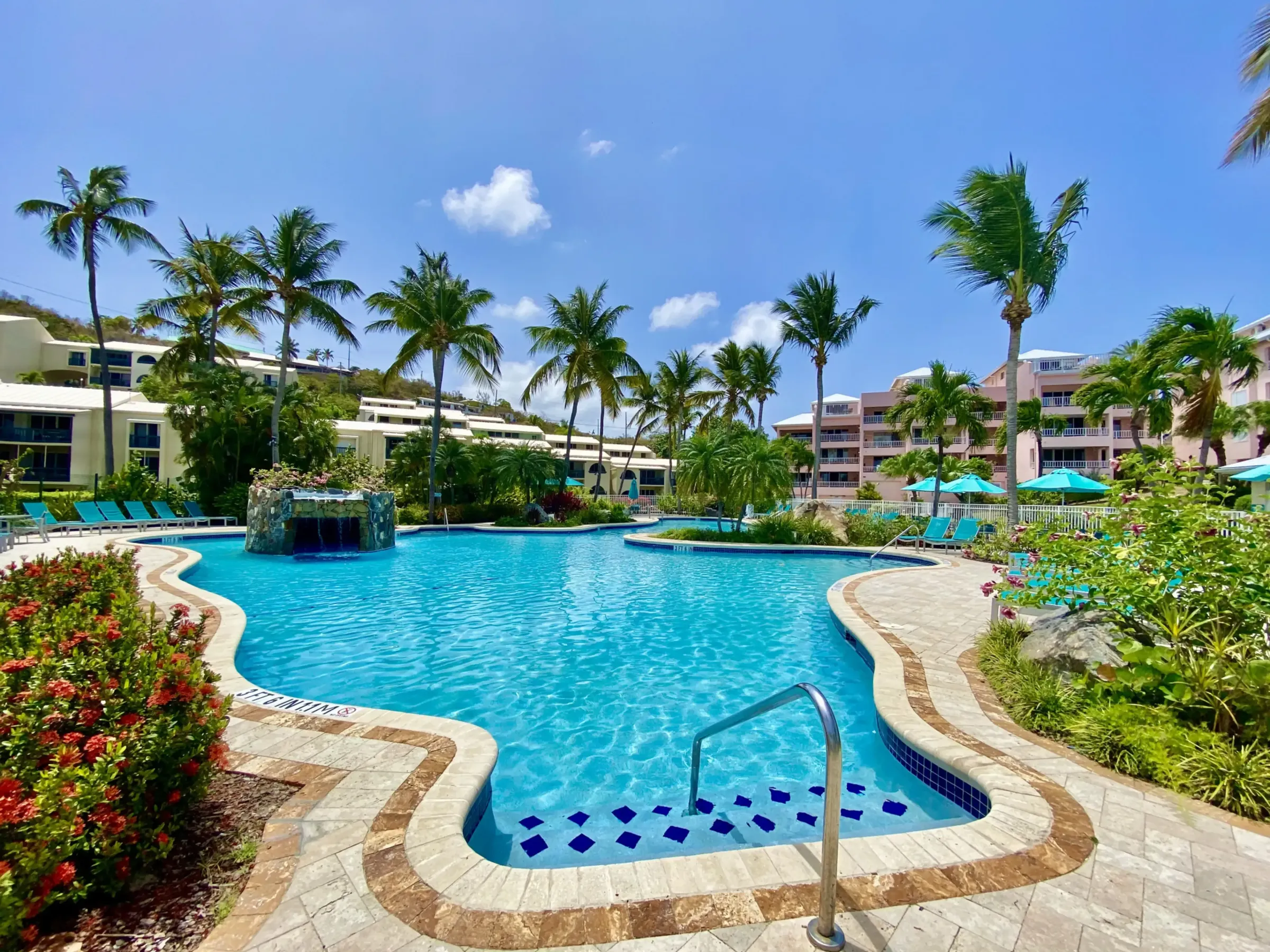 Tropical resort pool with palm trees, a waterfall, and lounge chairs under a bright blue sky.