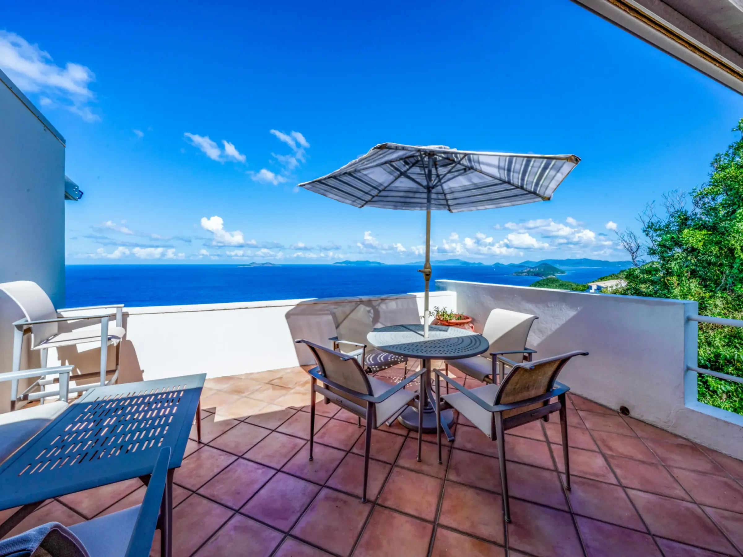 Terrace with table, chairs, and umbrella overlooking ocean and islands under a clear blue sky.