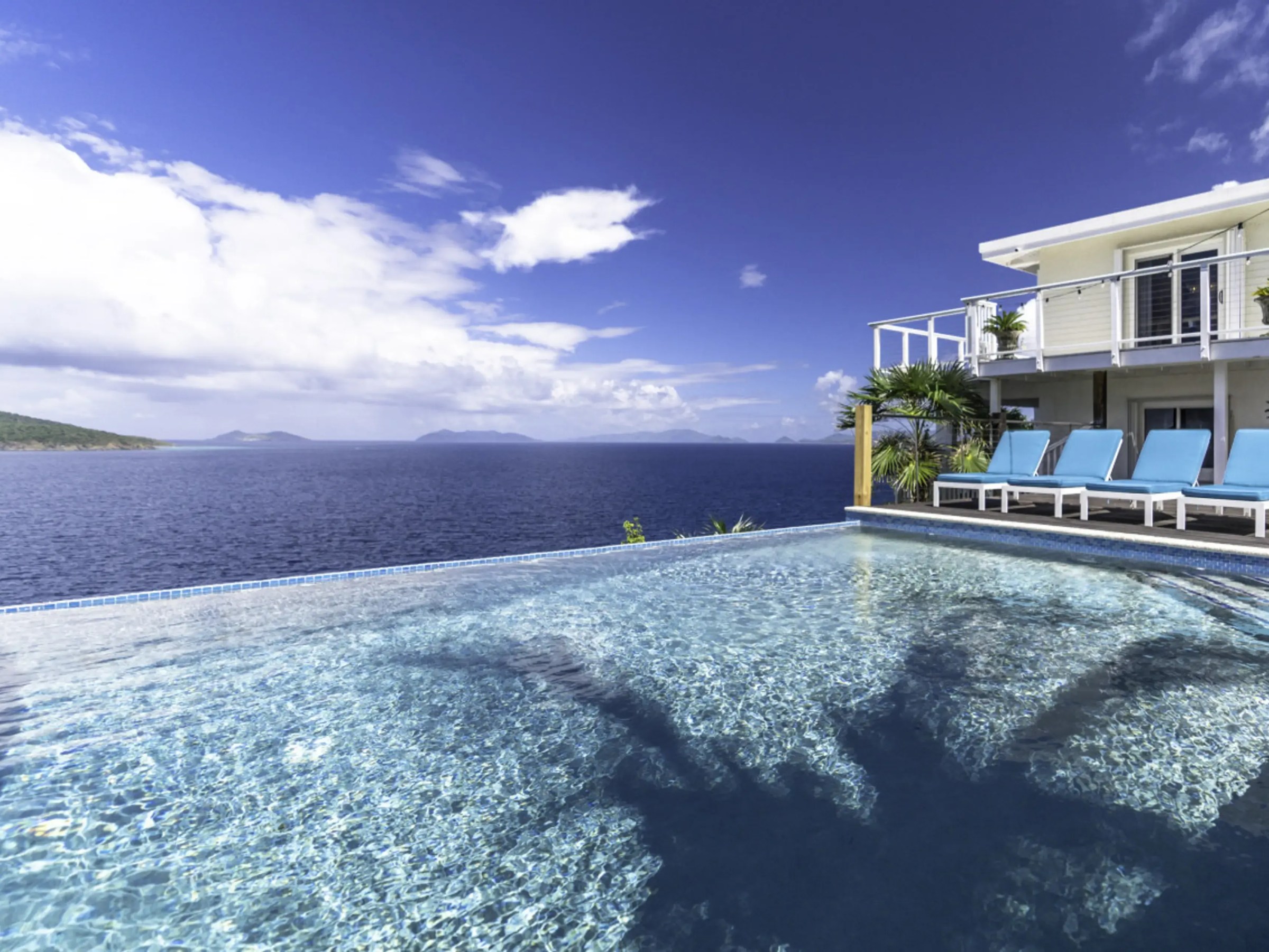 Infinity pool with sea view, lounge chairs, and modern house under a blue sky.