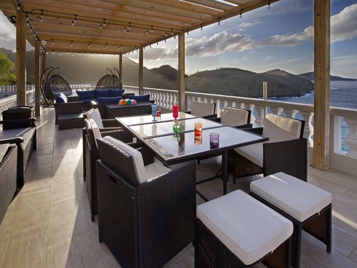Outdoor patio with wicker furniture, string lights, ocean view, and mountains at sunset.