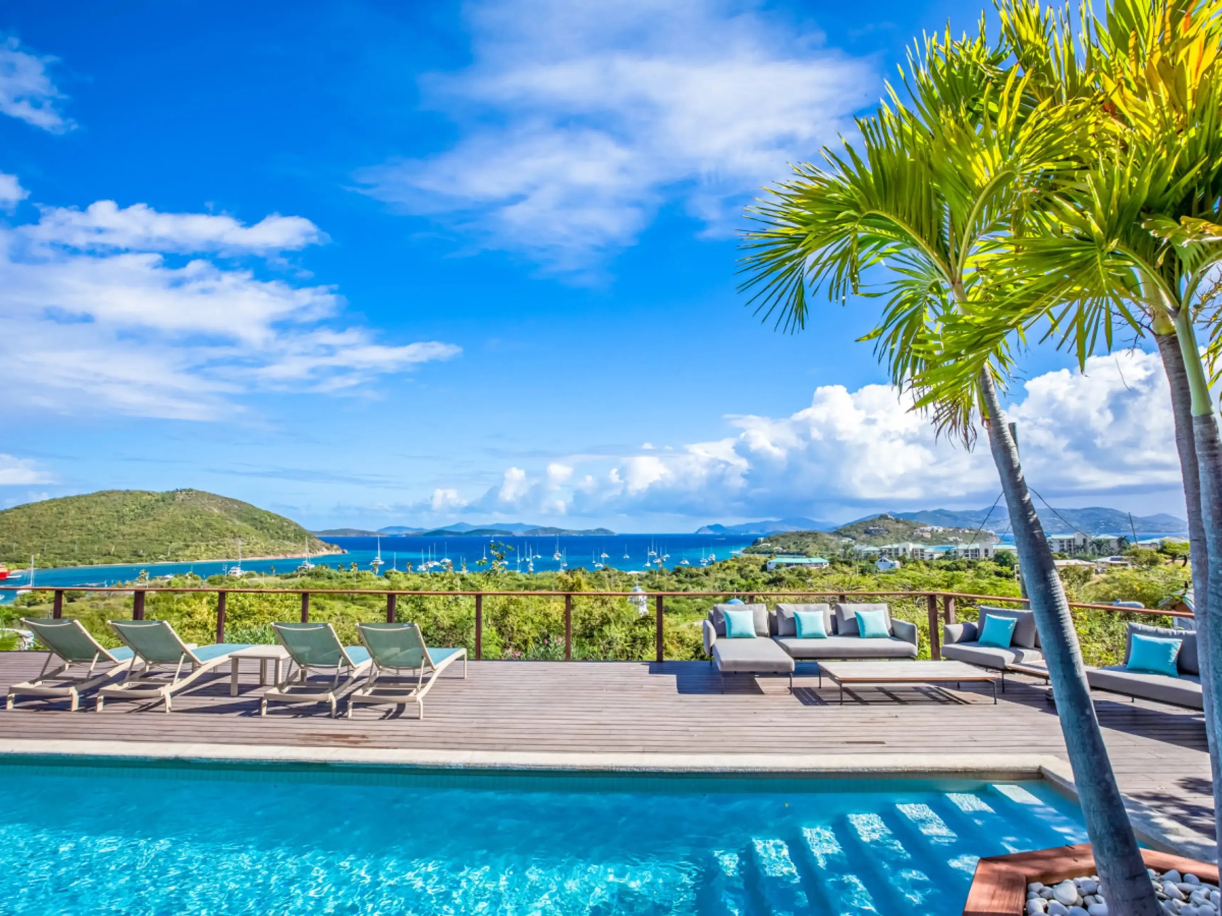 Poolside deck with lounge chairs, overlooking ocean and islands, under a blue sky with clouds and palm trees.