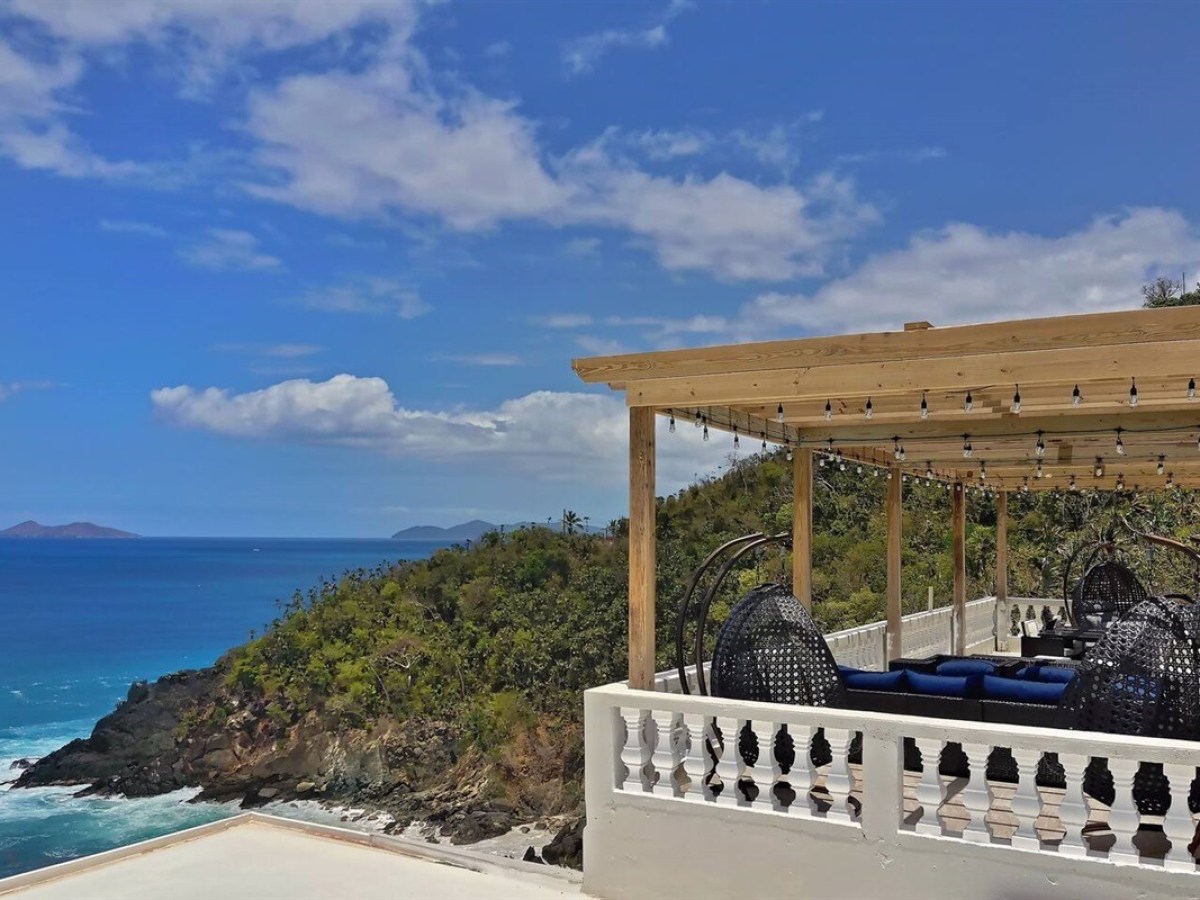 Coastal view from a terrace with wicker chairs under a wooden pergola, overlooking blue ocean and green hills.