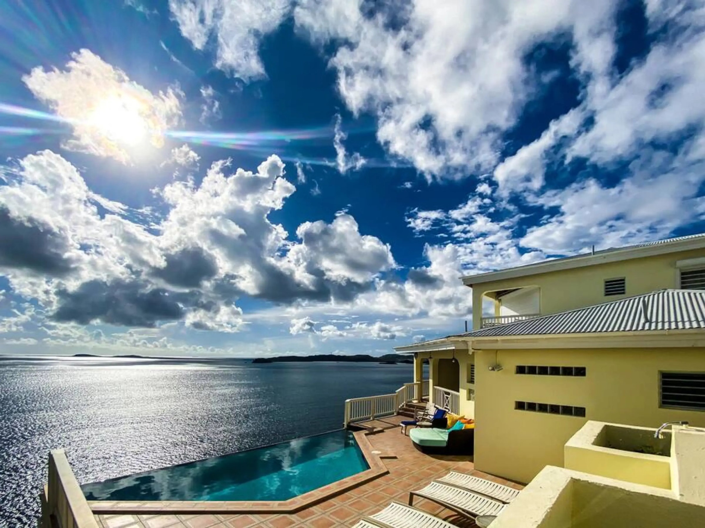 Sunny coastal view with clouds, a yellow house, and an infinity pool overlooking the ocean.
