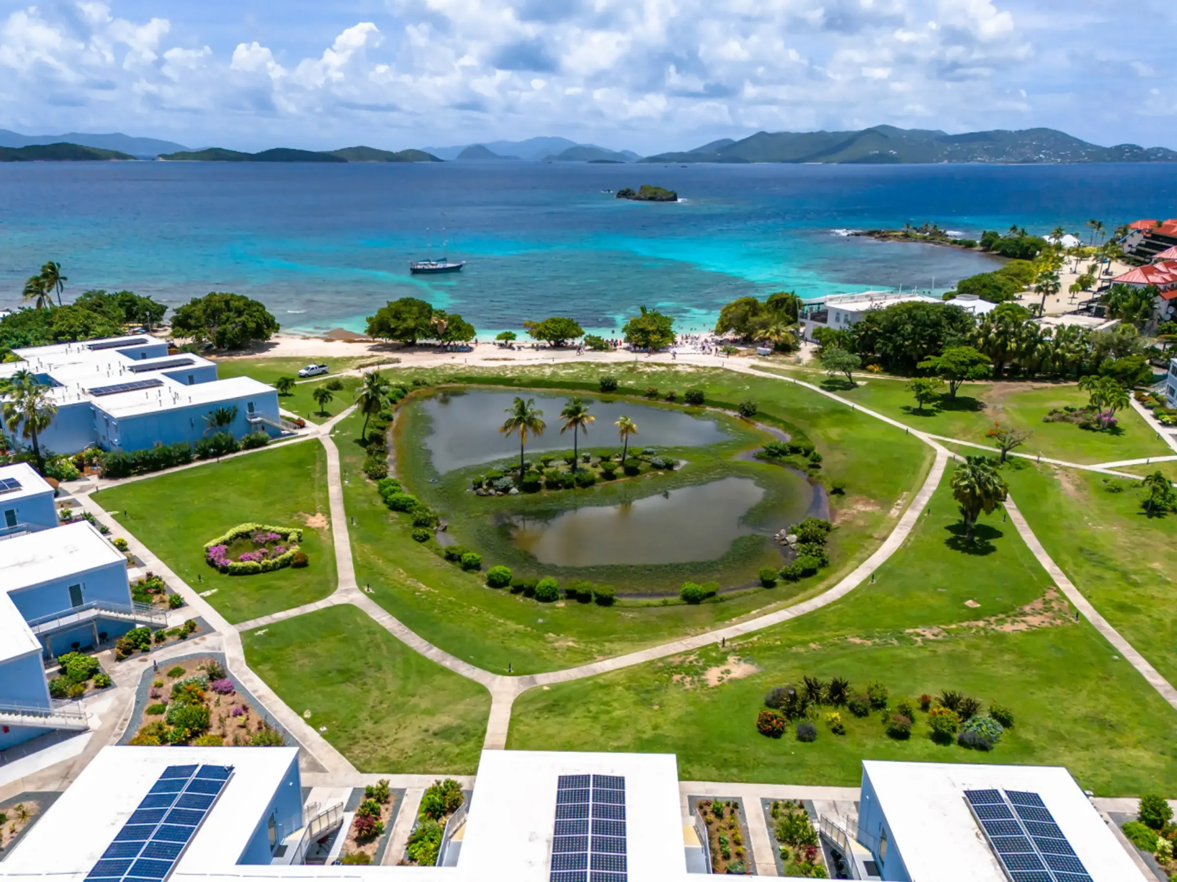 Aerial view of coastal resort with solar panels, pond, greenery, and ocean in background.