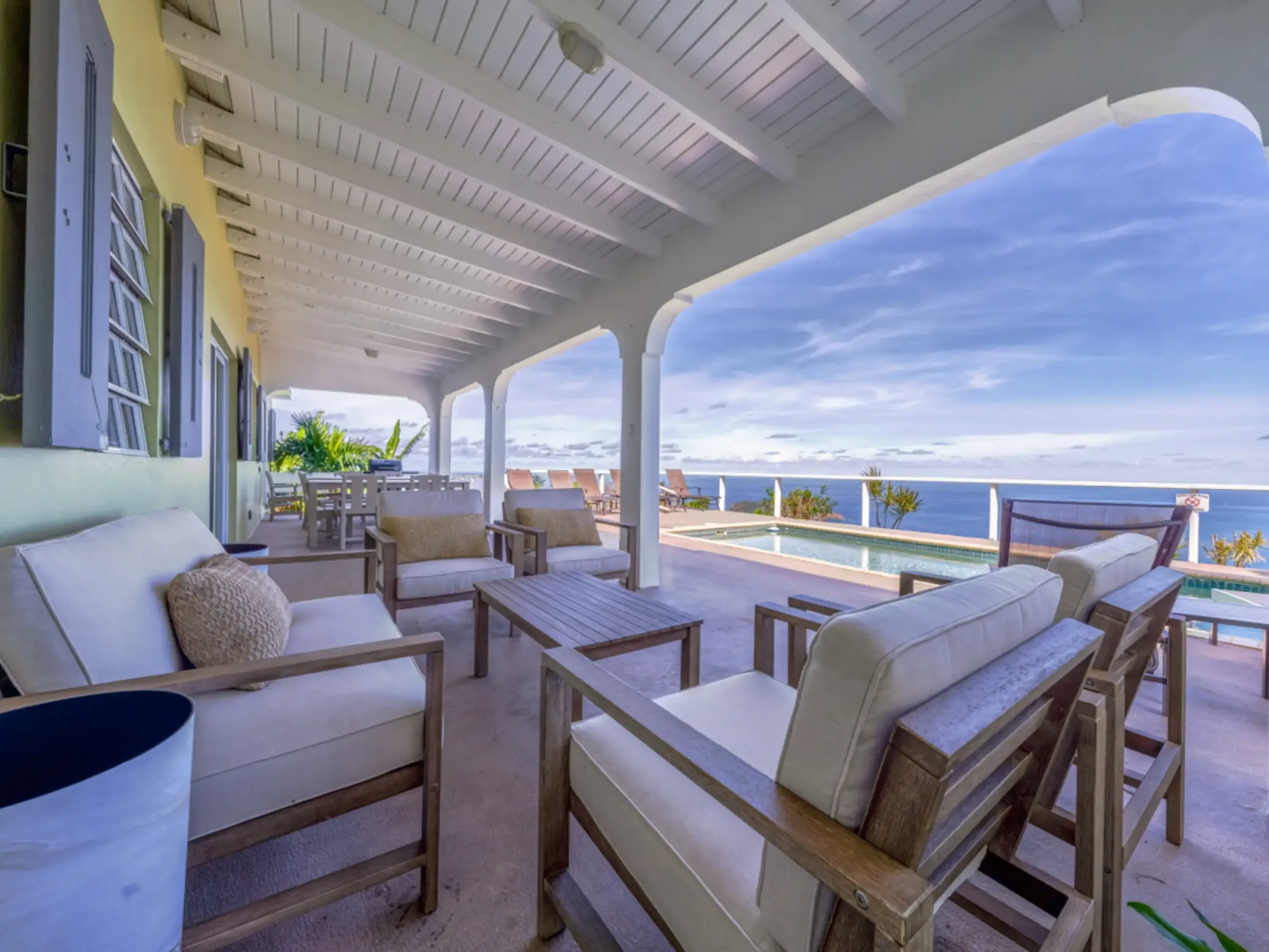 Covered patio with wooden furniture overlooking a pool and ocean view.