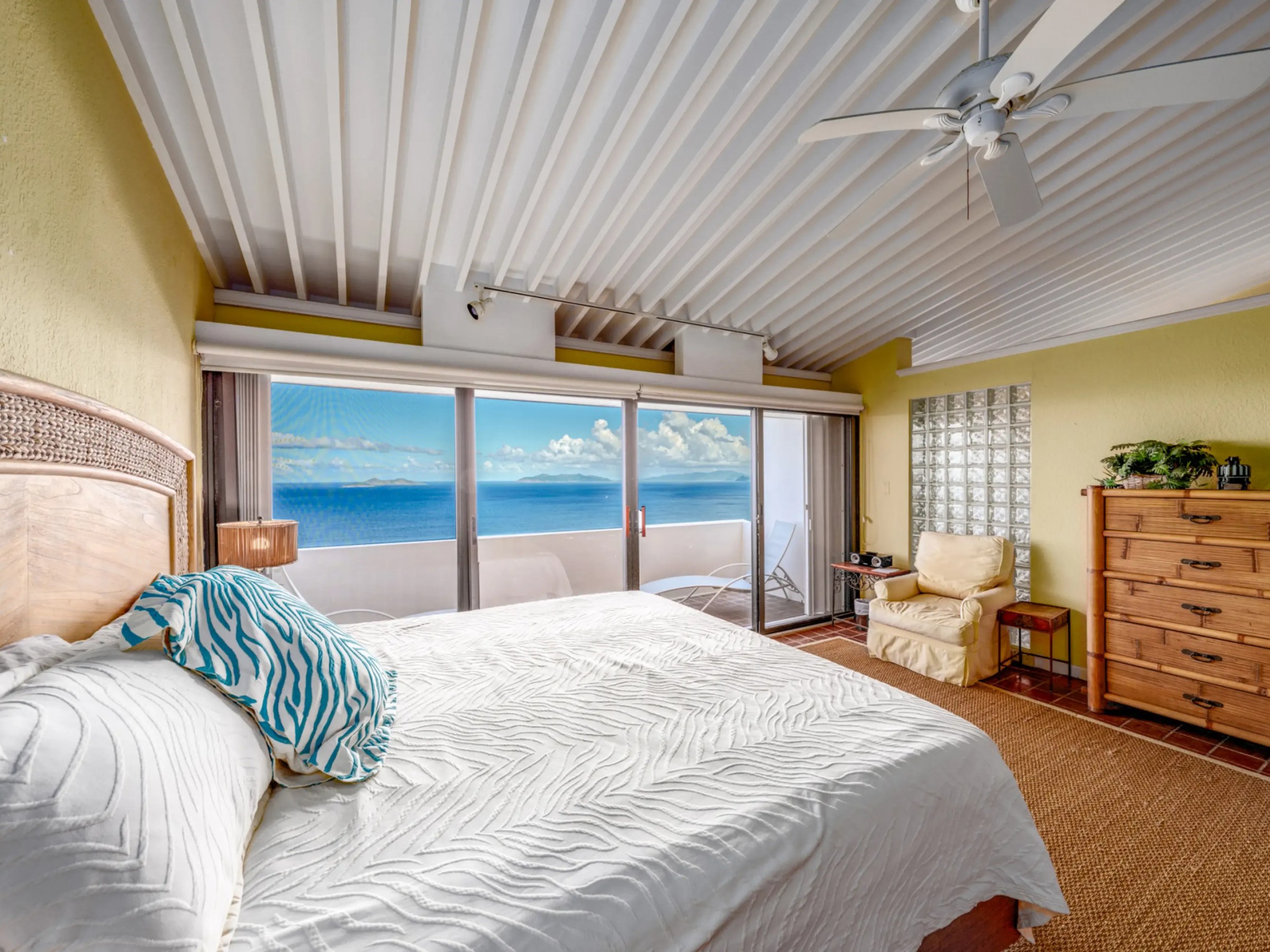 Bedroom with ocean view, large bed, ceiling fan, wooden dresser, and sliding glass doors to balcony.