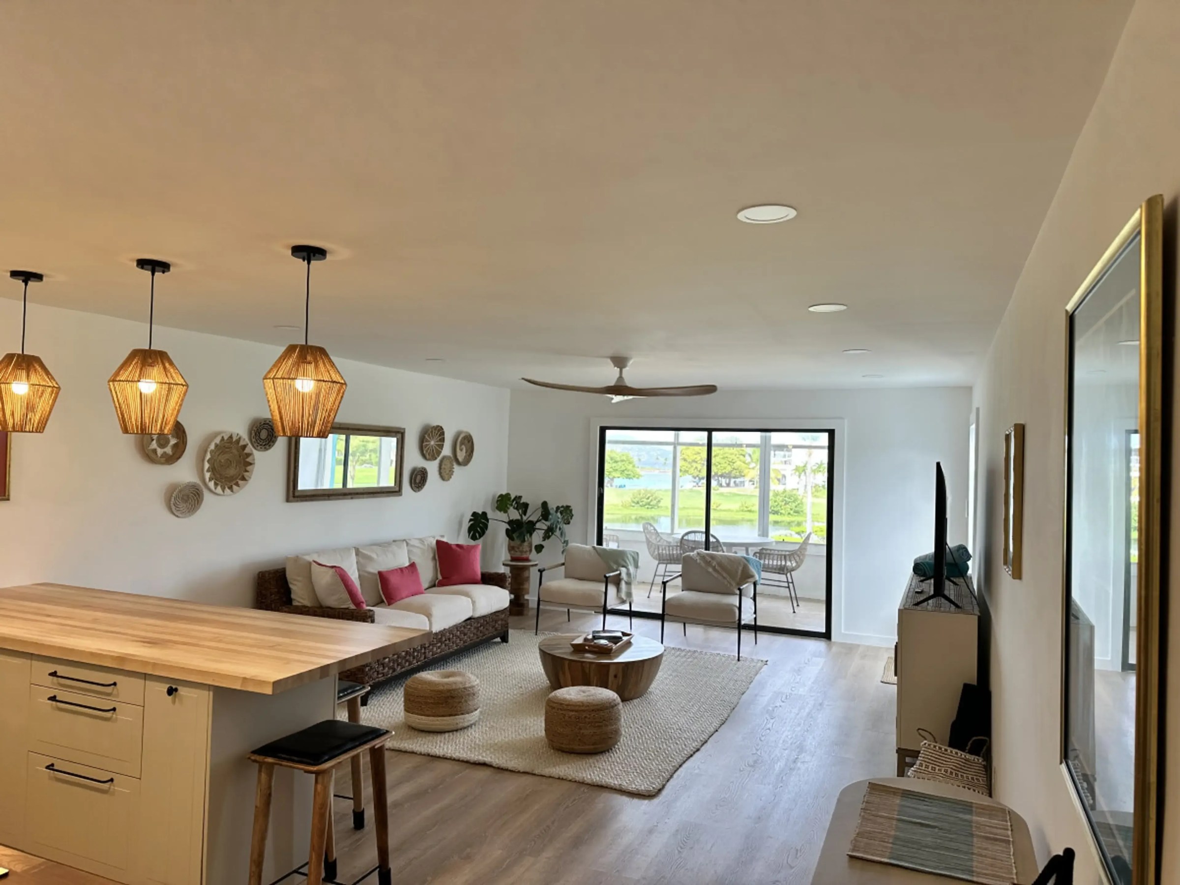 Open living room with wooden decor, rattan furniture, and large windows overlooking greenery.