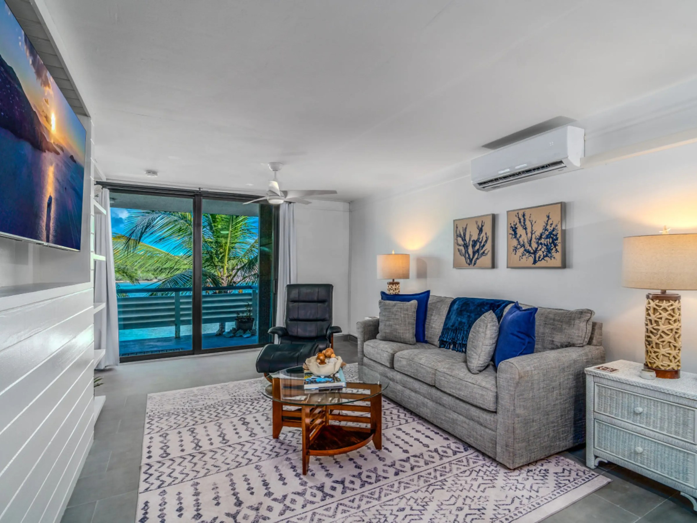 Living room with gray couch, blue pillows, tropical view, and TV on the wall.