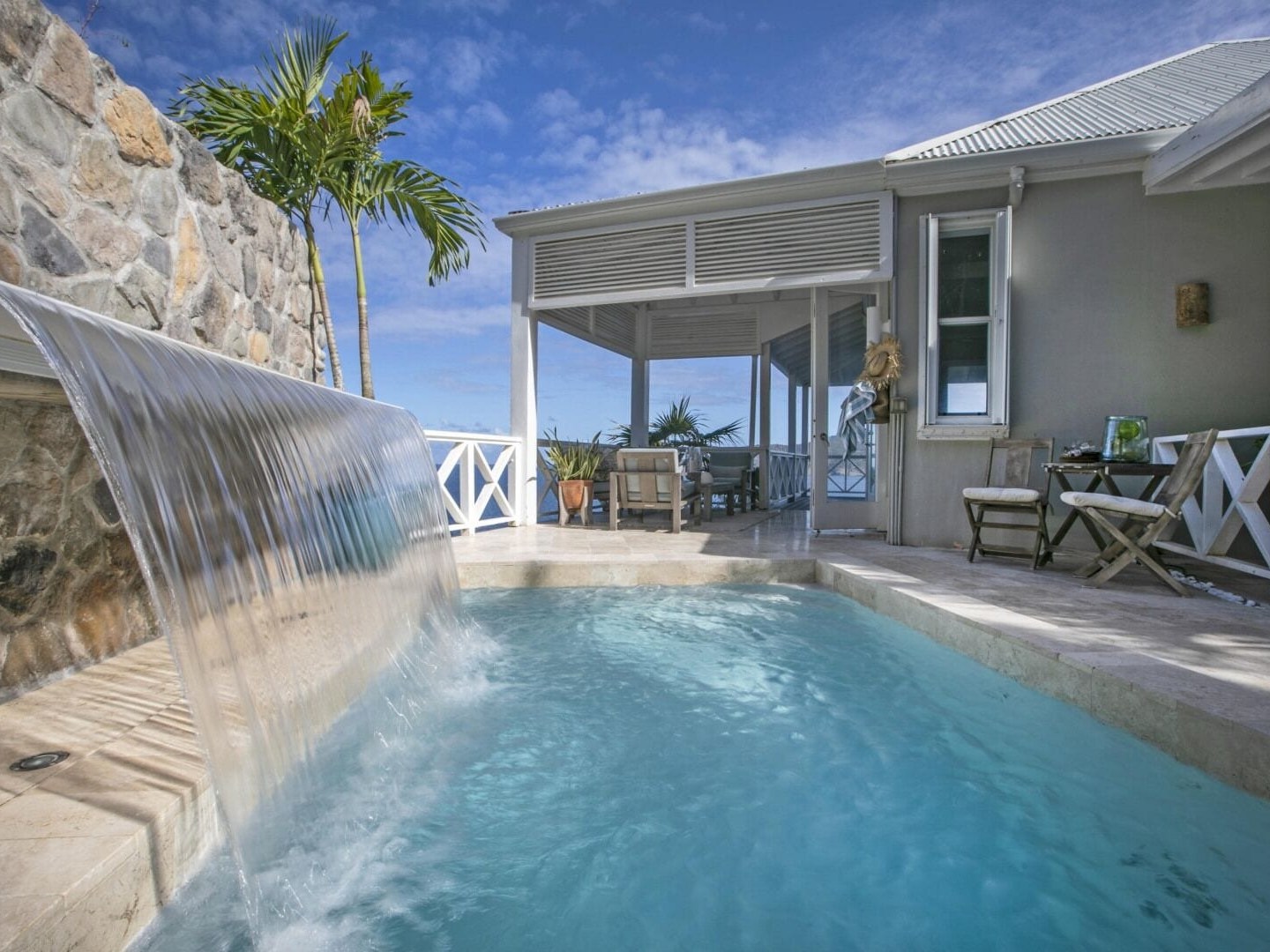 A pool with a waterfall feature beside a patio and stone wall under a blue sky.