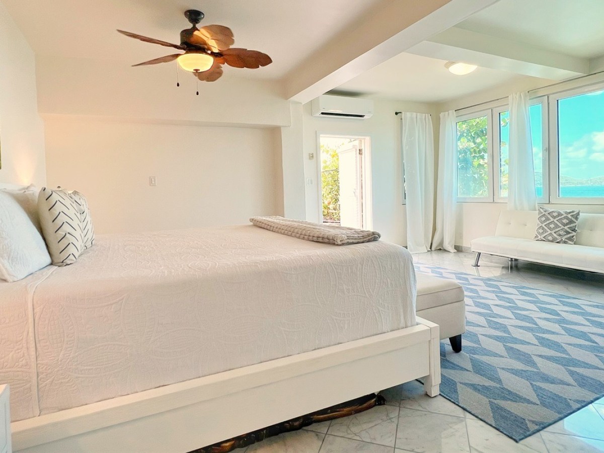 Bright bedroom with white bed, ceiling fan, couch, rug, and ocean view through large windows.
