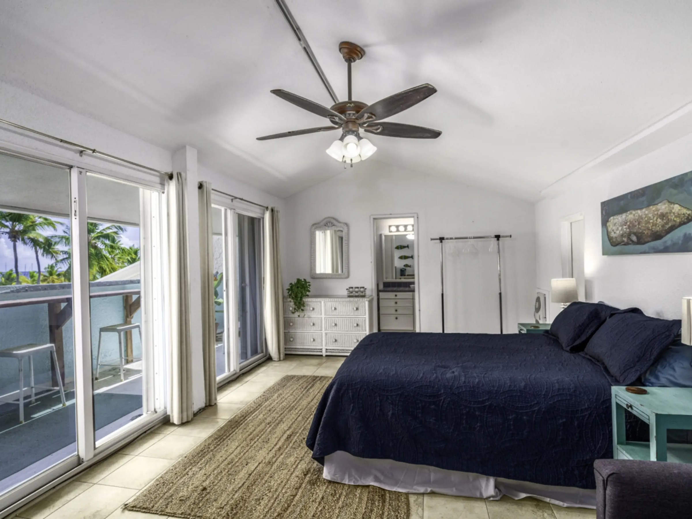 Spacious bedroom with dark blue bed, ceiling fan, balcony view, and sliding glass doors.