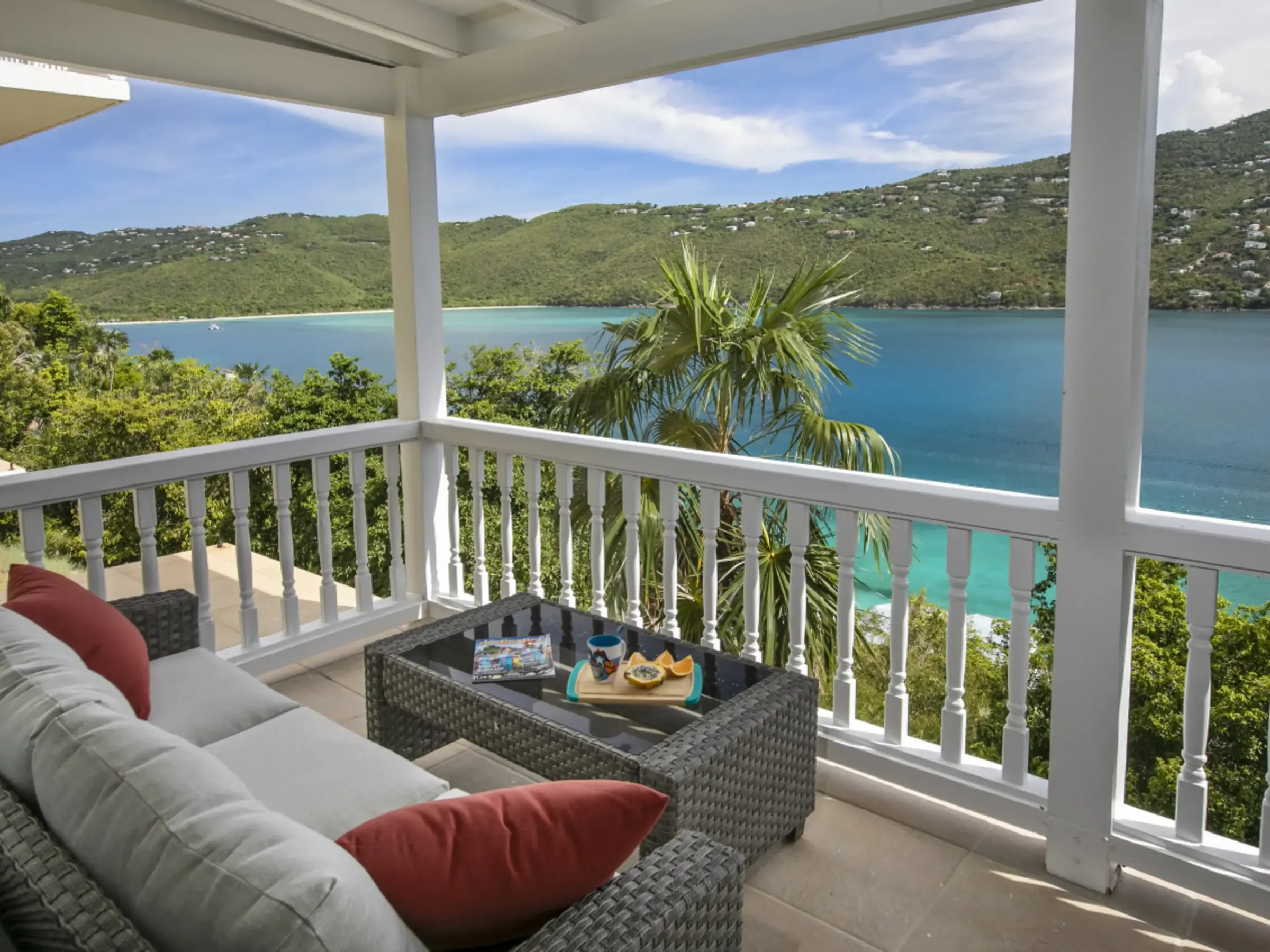 Covered balcony view of a bay with a wicker sofa and table, and hills in the background.