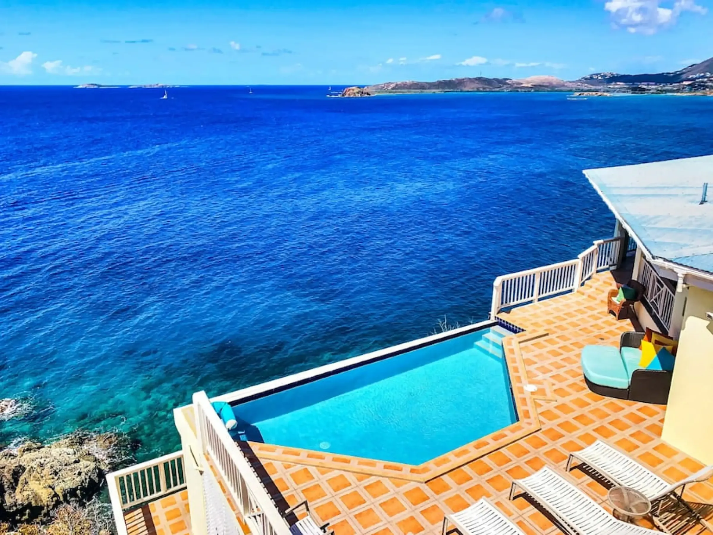 Ocean view with a pool and sun loungers on a tiled terrace next to a building.