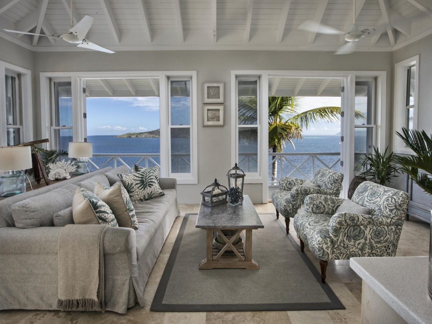 Coastal living room with ocean view, gray sofa, two patterned chairs, and coffee table with lanterns.