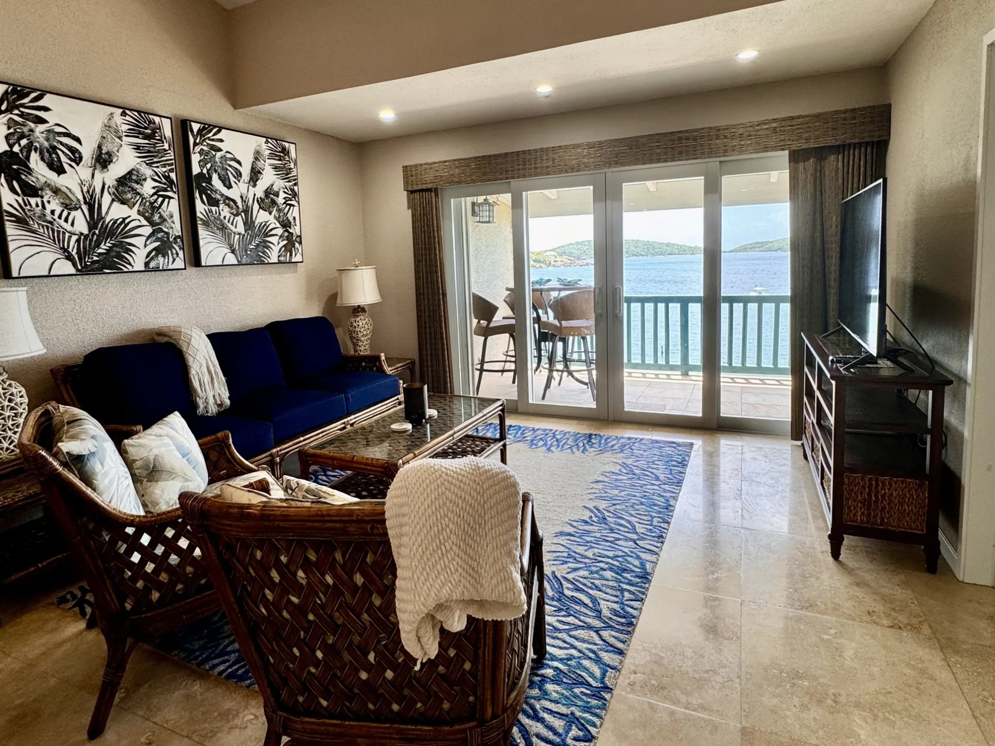 Living room with blue sofa, patterned rug, tropical art, and doors opening to a balcony with sea view.