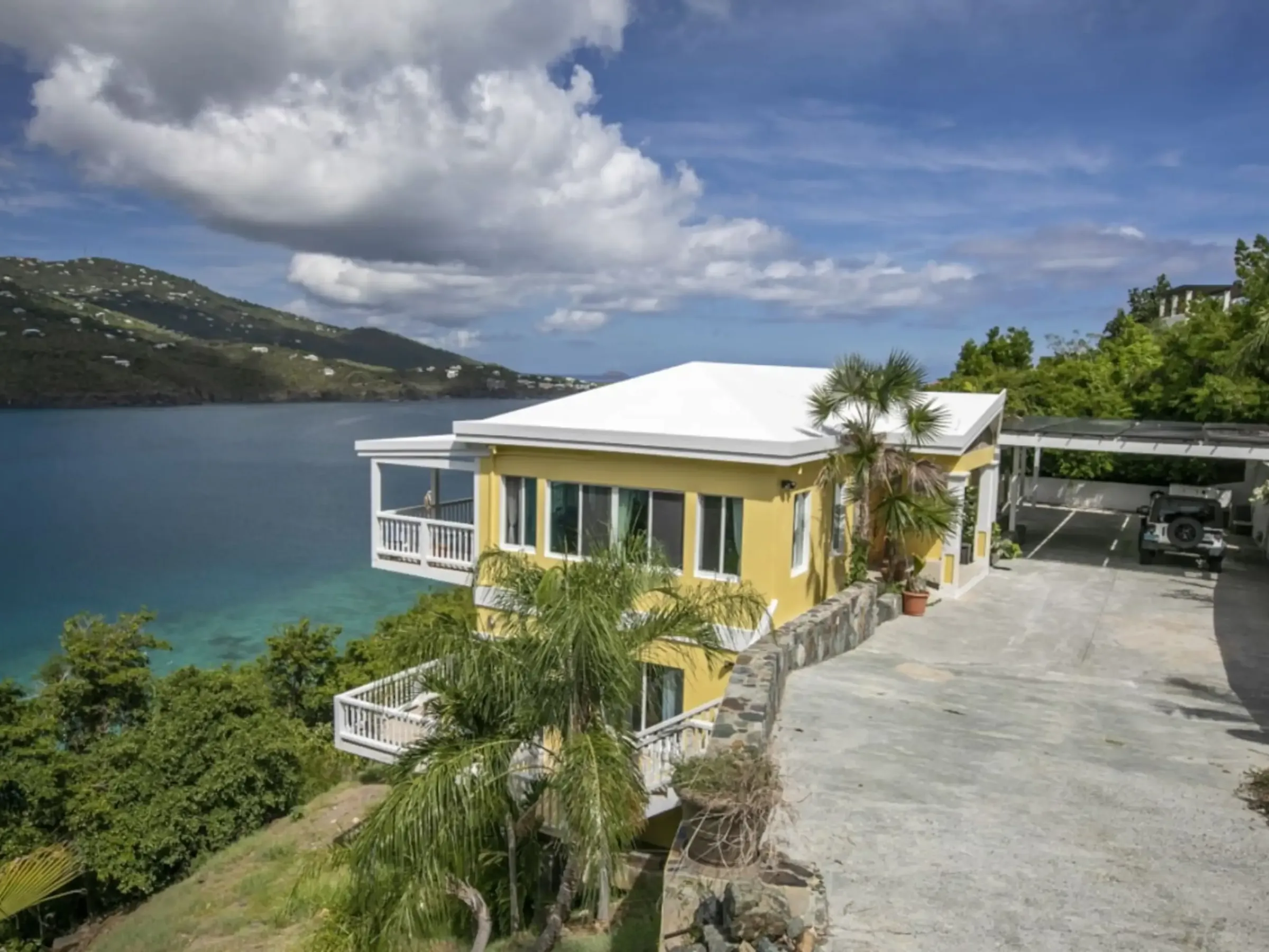 Seaside yellow house with white roof, overlooking blue water, surrounded by palm trees under a cloudy sky.